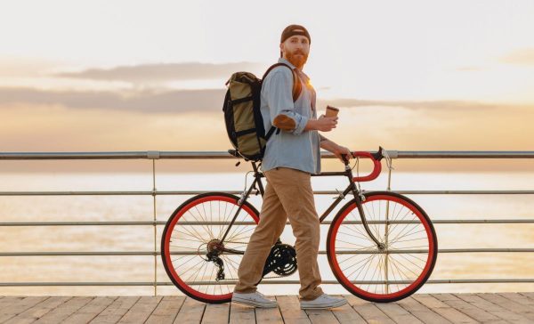 handsome hipster style bearded man with backpack wearing denim shirt and cap with bicycle in morning sunrise by the sea drinking coffee, healthy active lifestyle traveler backpacker