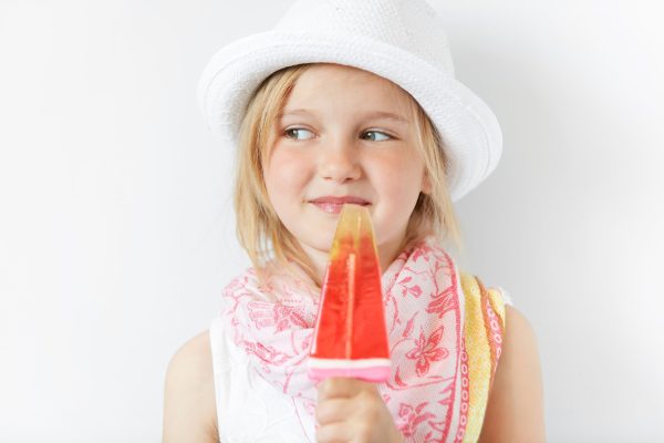 Smiling child with popsicle cunningly looking sideways indoors. Positive emotions, contended look and summer careless atmosphere makes kid look nice and shinny.